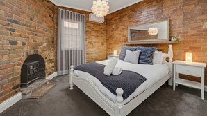 Een slaapkamer met een bakstenen muur, een open haard, een bed met wit hoofdeinde, een nachtkastje en een hangende lamp.