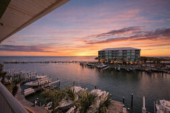 A marina with boats docked, a building with multiple floors, and a sunset view. - Beach N Bay 2 BR Navy Cove Condo w\ FREE Boat Slip (Gulf Shores)