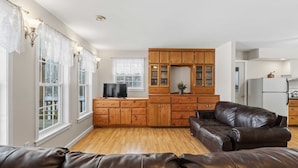 A living room with a brown leather sofa, wooden cabinets, a television, and a kitchen area with a refrigerator. - Storybook Craftsman Cottage - Annapolis Valley (Aylesford)