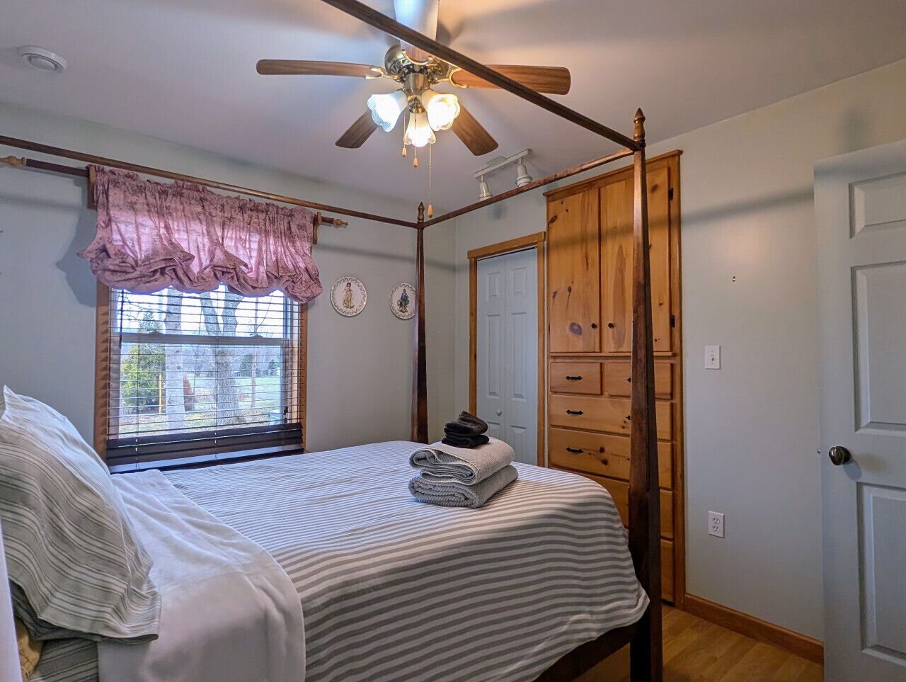 A bedroom with a bed, a ceiling fan, a window with blinds, a wooden wardrobe, and a door.