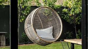 A wicker hanging chair with a striped cushion in a backyard with a wooden table and a metal chair.