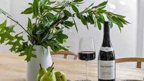 A dining table with a white vase, green pears, a bottle of wine, and a plate with cheese.