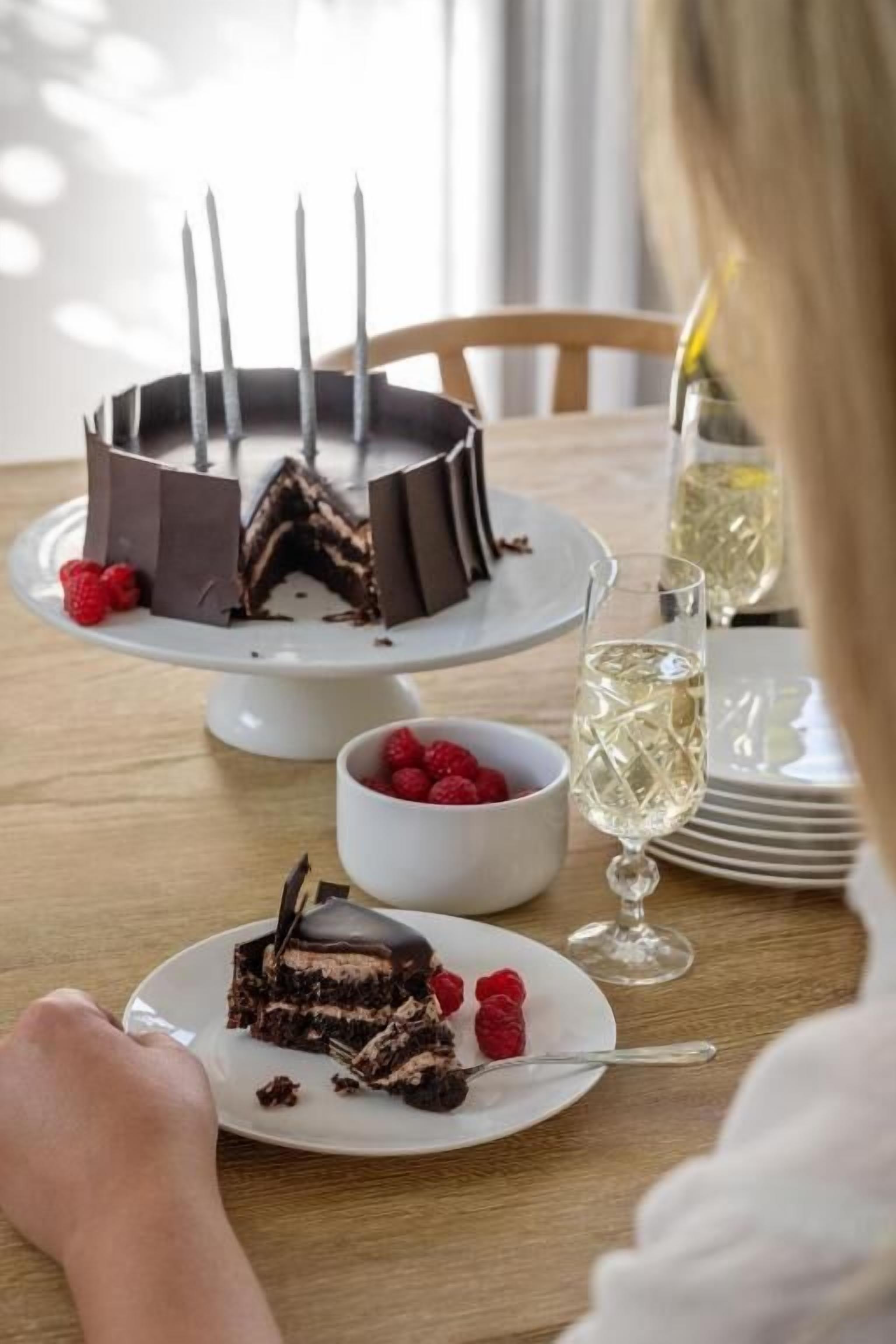 A cake with a slice cut out on a white plate, a bowl of raspberries, and a glass of champagne on a wooden table.