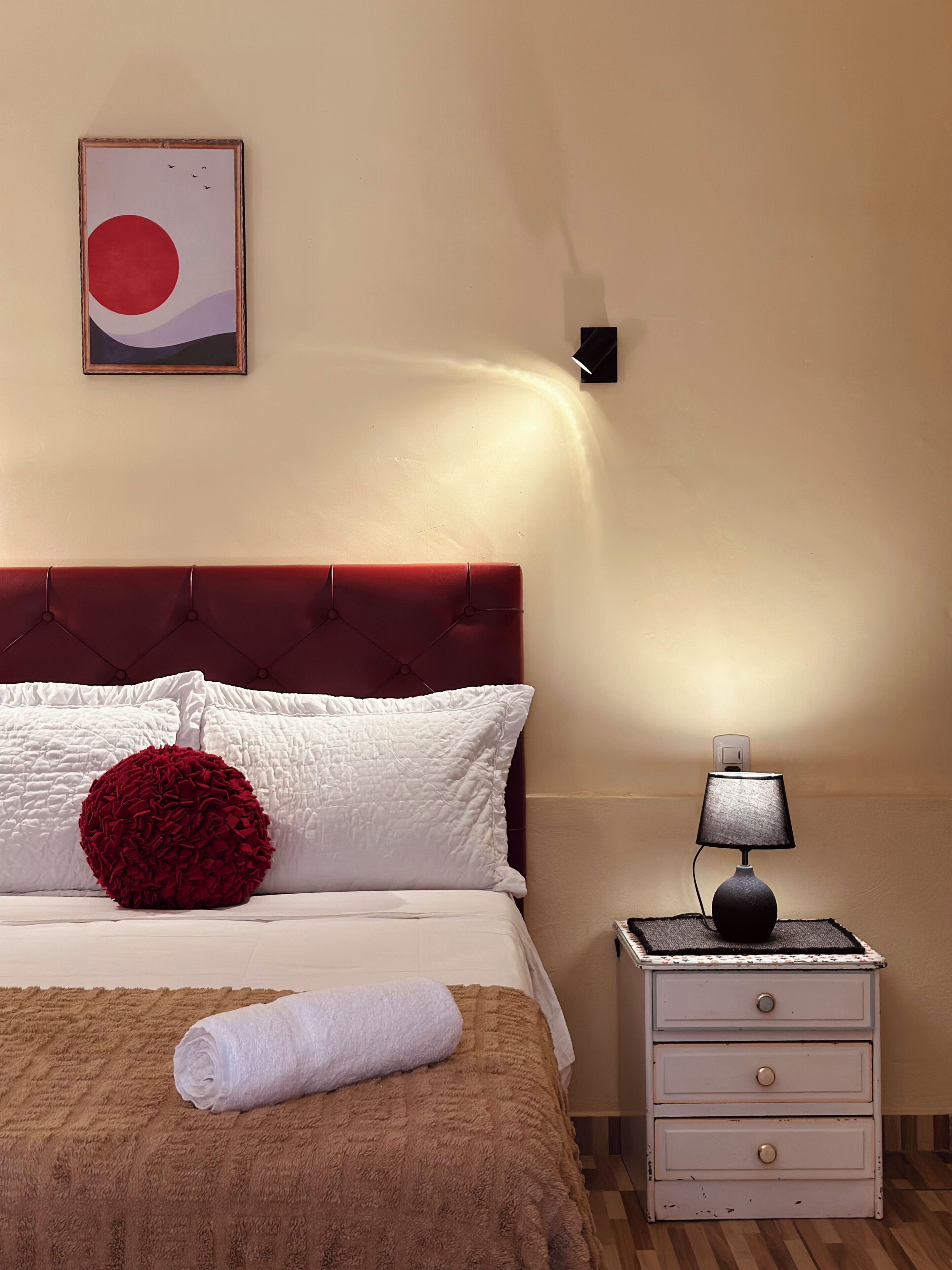 Een netjes opgemaakt bed met een rood kussen, een witte deken en een bruine plaid. Er staat een klein nachtkastje met een lamp en een lade. Aan de muur boven het bed hangt een ingelijste foto.