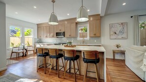 Une cuisine moderne avec des meubles en bois, un îlot central doté de tabourets de bar, un éclairage suspendu et une vue sur l’extérieur.