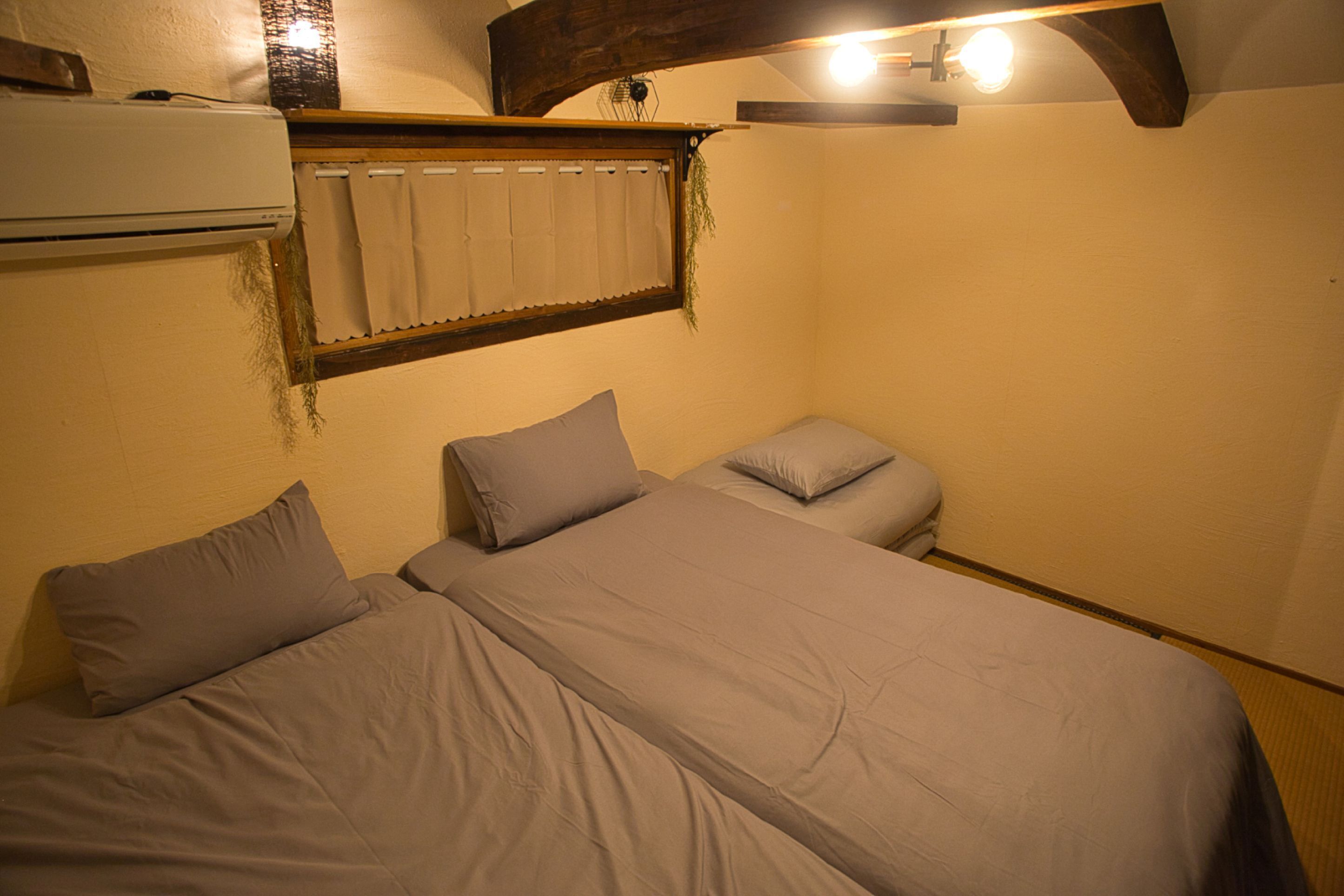 A room with two beds, a window with curtains, an air conditioning unit, and a wooden ceiling beam.
