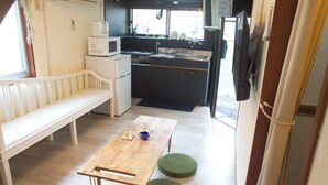 Cocina compacta con microondas, estufa, pileta y una televisión de pantalla plana. Hay una mesa de madera con dos taburetes verdes y un banco blanco en un pequeño estar.