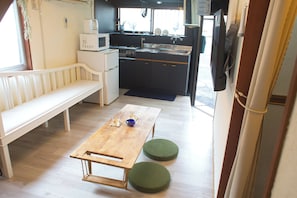 A compact kitchen with a microwave, stove, sink, and a flat-screen TV. There's a wooden table with two green stools and a white bench in a small living area.