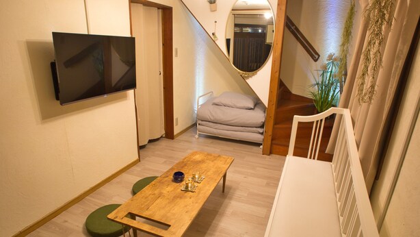 Una habitación con una mesa de madera, un taburete con cojín verde, un radiador blanco, un televisor montado en la pared y una escalera con una planta.