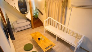 Una habitación pequeña y acogedora con una mesa de madera, dos taburetes verdes, un banco blanco, un televisor y una escalera que lleva a otra zona.