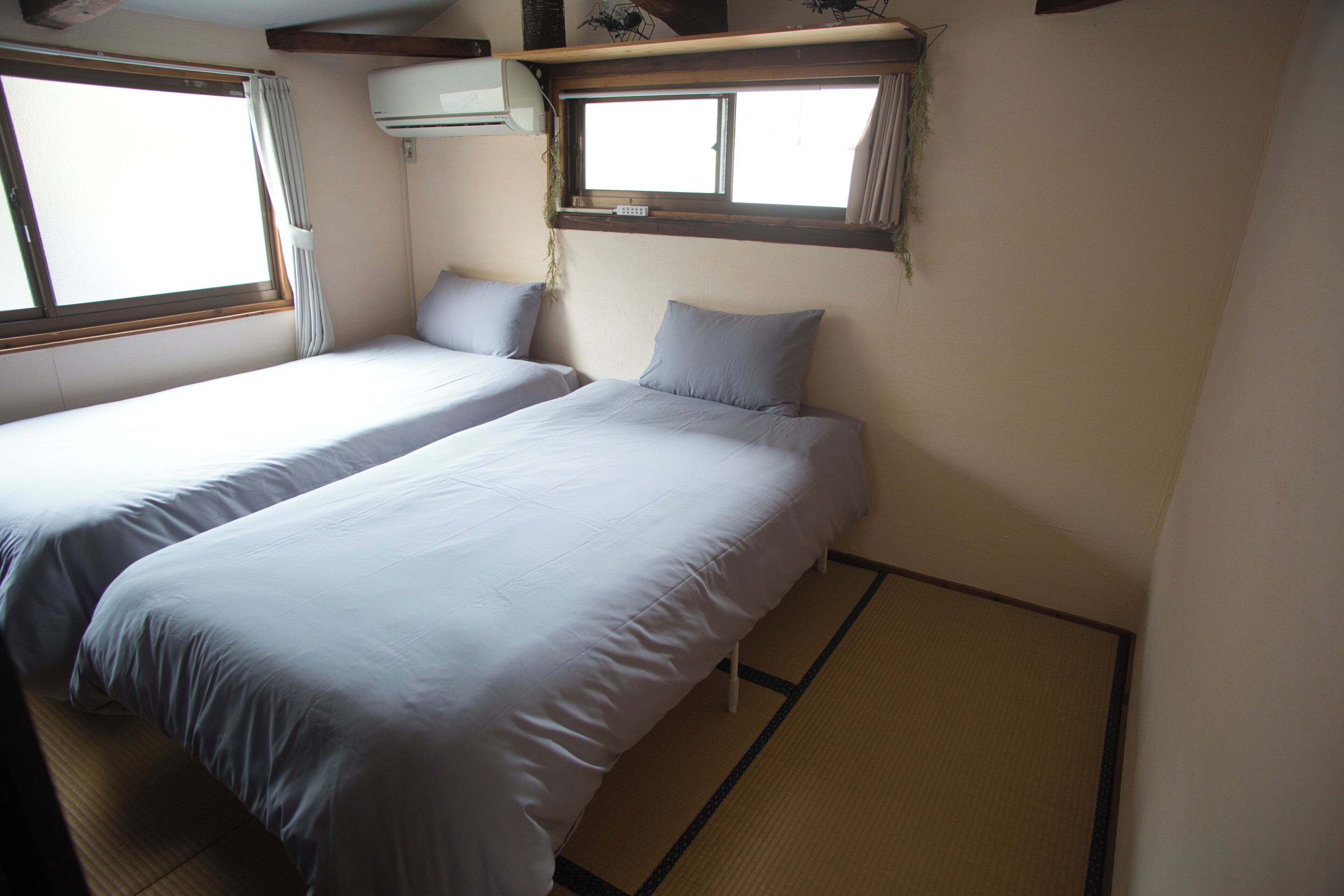Een kamer met twee bedden, een raam, een airconditioner en een klein plankje met spullen.