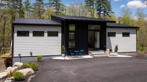 A modern cabin with a black roof, white siding, and a covered porch with blue chairs. - Brand new riverfront home with private beach, modern, serene, central HV spot. (High Falls)