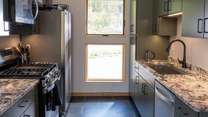 A compact kitchen with a gas stove, stainless steel appliances, and a window overlooking a natural landscape. - Brand new riverfront home with private beach, modern, serene, central HV spot. (High Falls)