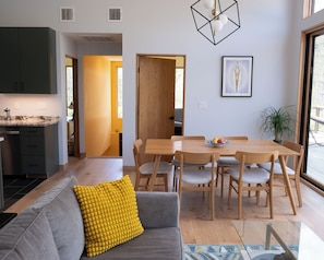 A modern kitchen with dark cabinets, a dining area with a wooden table and chairs, and a bright yellow cushion on a grey sofa.