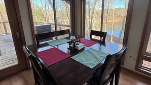 Comedor con mesa de madera, cuatro sillas y vistas a árboles y un cuerpo de agua a través de amplias ventanas.