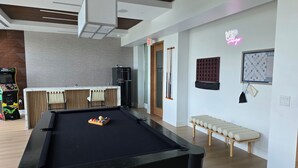 A pool table in a modern room with a bar area, a neon sign, and a seating bench. - Lux in Black 2BR Bay, Pool & Ocean Views (Fort Myers)