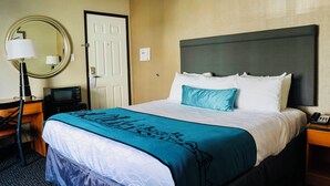 Une chambre d’hôtel avec un lit, un bureau, une chaise, une lampe, un miroir et une porte.