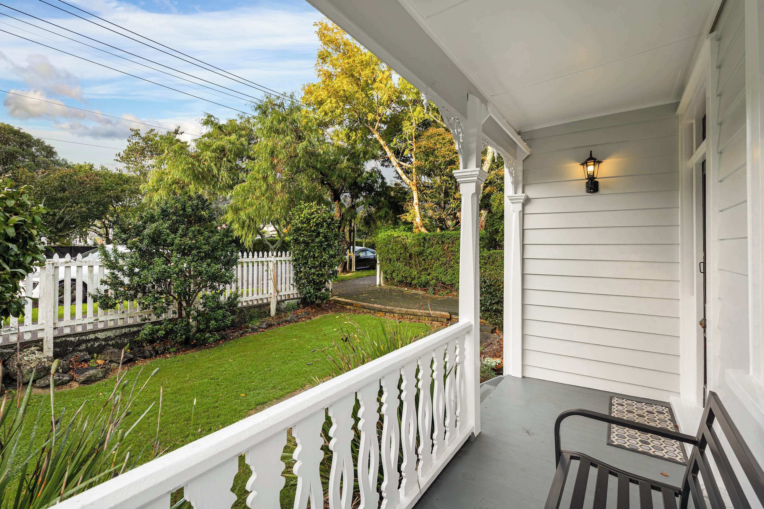 A porch with a white railing, a bench, and a street view with a white fence and trees.