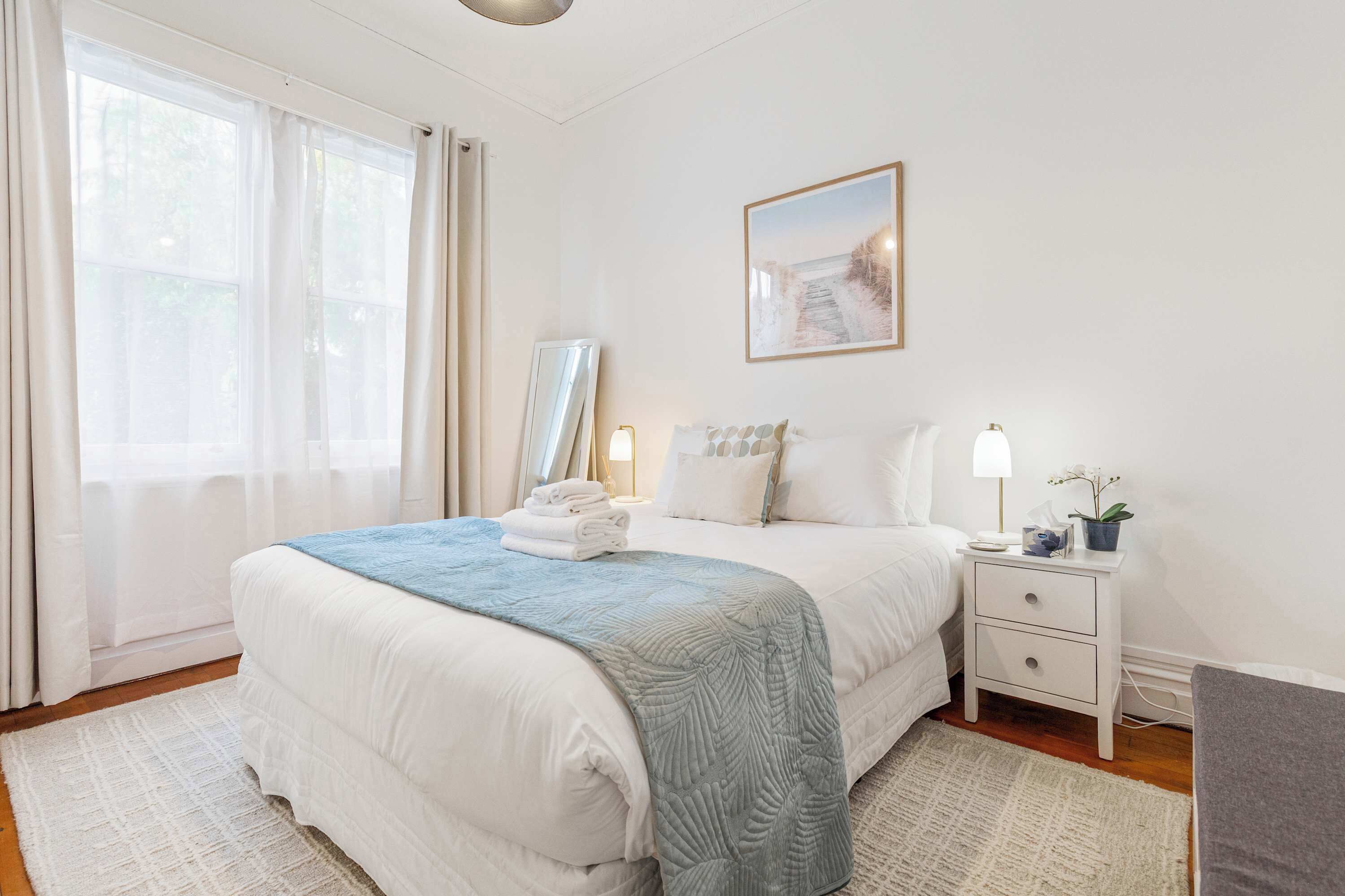 A neatly arranged bedroom with a bed, bedside tables, a nightstand, a framed picture, and a window with curtains.