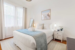 Ein ordentlich eingerichtetes Schlafzimmer mit einem Bett, Nachttischen, einem Fenster mit Vorhängen und einem gerahmten Bild.