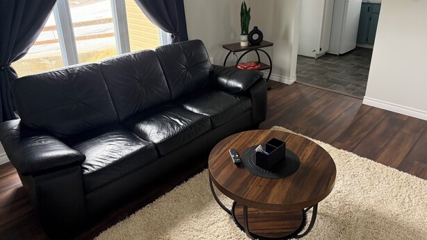Una sala de estar con un sofá de cuero negro, una mesa de centro redonda de madera y una cocina al fondo.