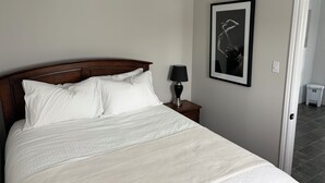 Una cama bien hecha con sábanas blancas, un cabecero de madera oscura, una lámpara de noche y un cuadro enmarcado en la pared.