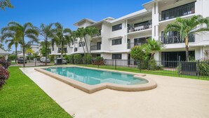 Un edificio moderno de varios pisos con piscina, rodeado de céspedes cuidados y palmeras.