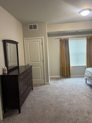 A bedroom with a dresser, a door, a window with curtains, and a ceiling light.