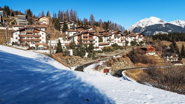 Un poble de muntanya cobert de neu, amb edificis de diversos pisos, un camí sinuós i una pendent nevada.