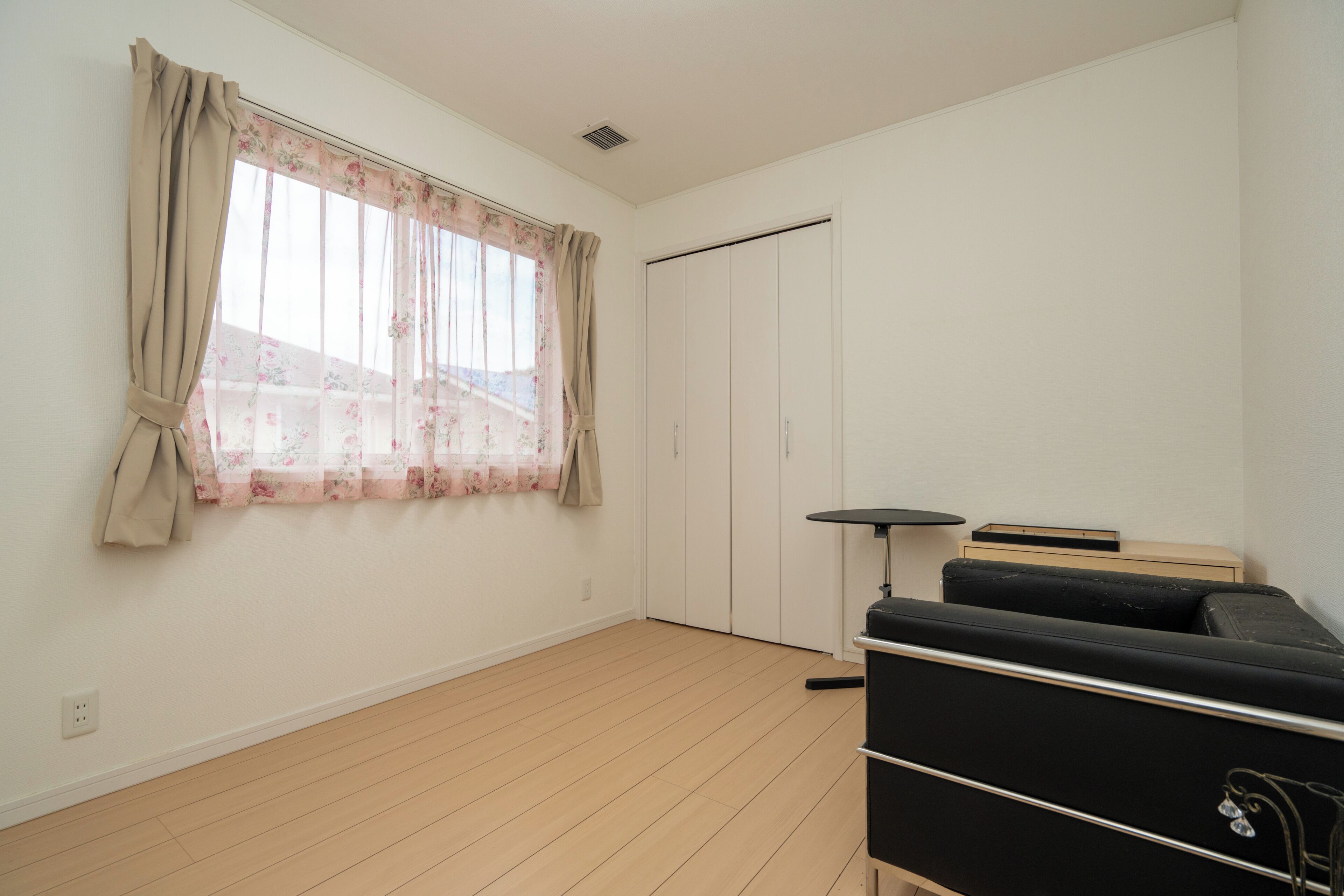 A room with a wooden floor, a black sofa, a small table, a wardrobe, and a window with floral curtains.
