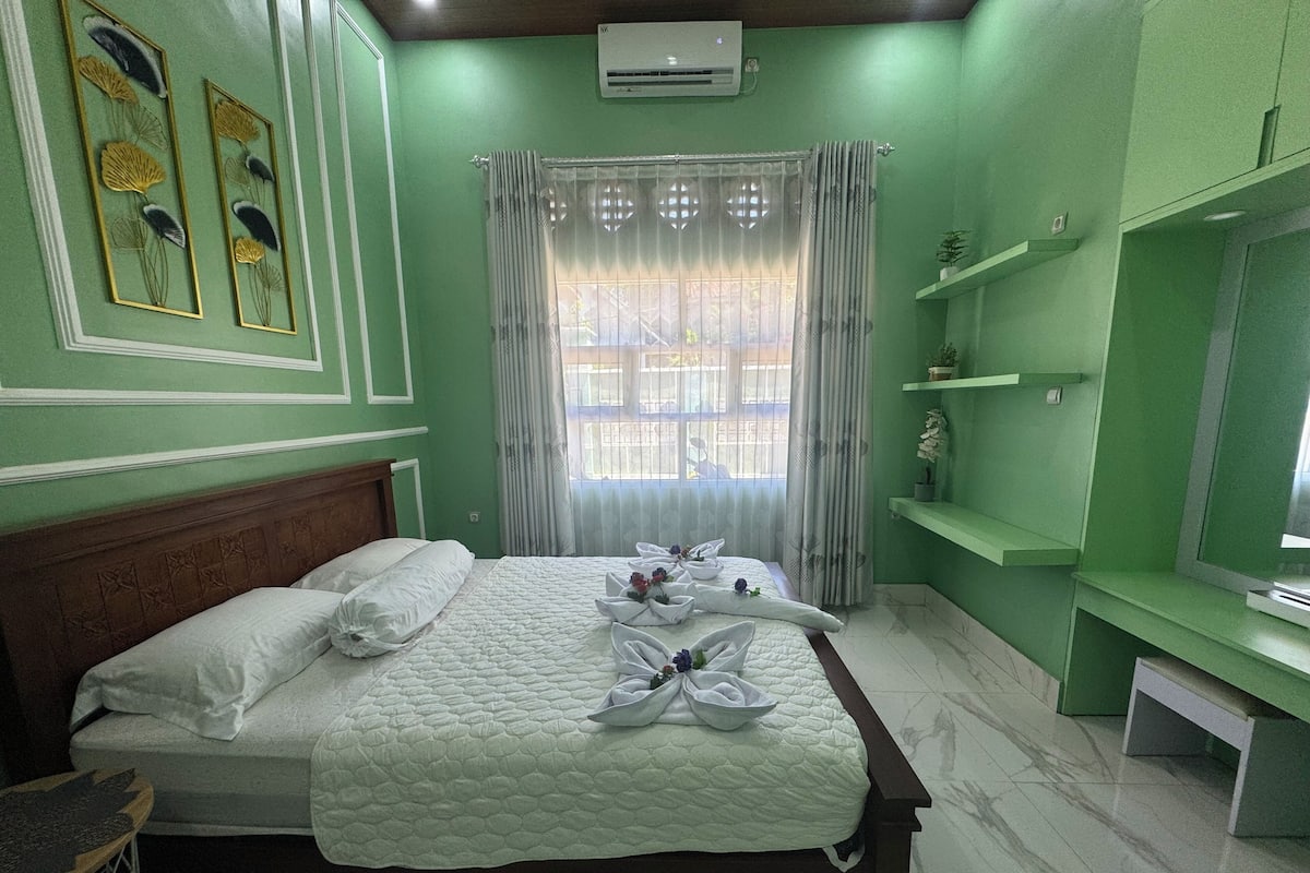 Un lit bien fait, agrémenté d’oreillers et de serviettes, une table de chevet, une fenêtre avec des rideaux et un mur vert orné d’éléments décoratifs.