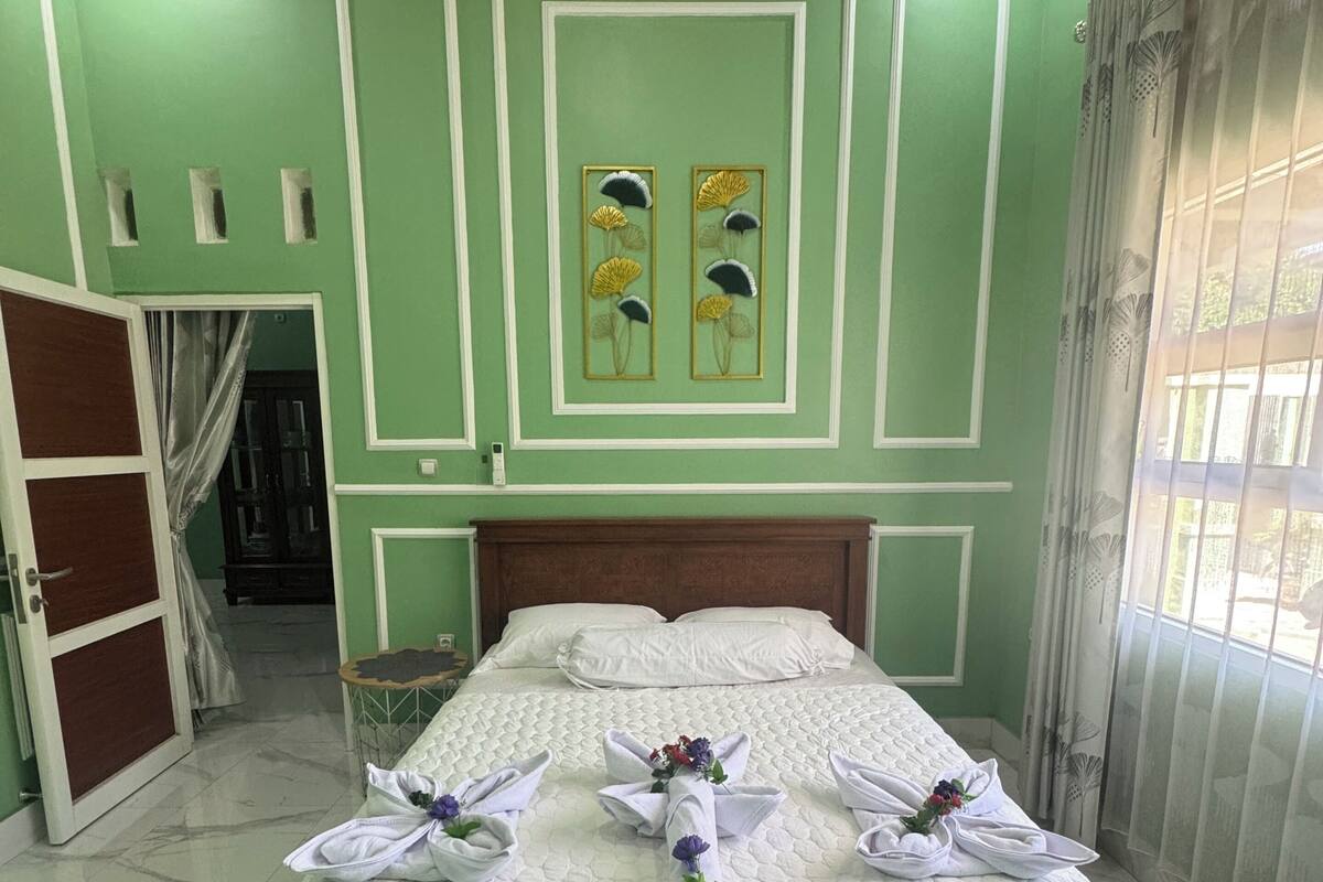 Un lit bien fait, agrémenté de serviettes et de fleurs, avec une tête de lit en bois et un mur vert orné de panneaux décoratifs.