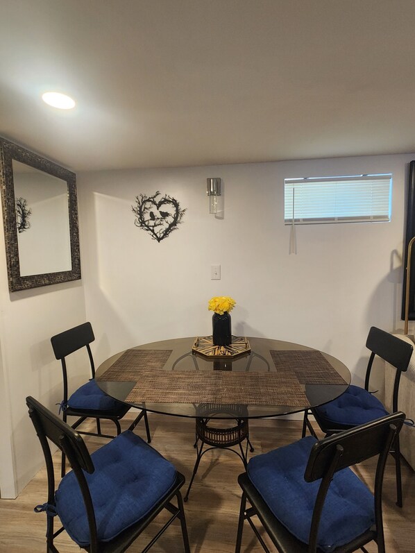 A dining area with a round table, four chairs, a vase with yellow flowers, a wall-mounted light, and a window with blinds.