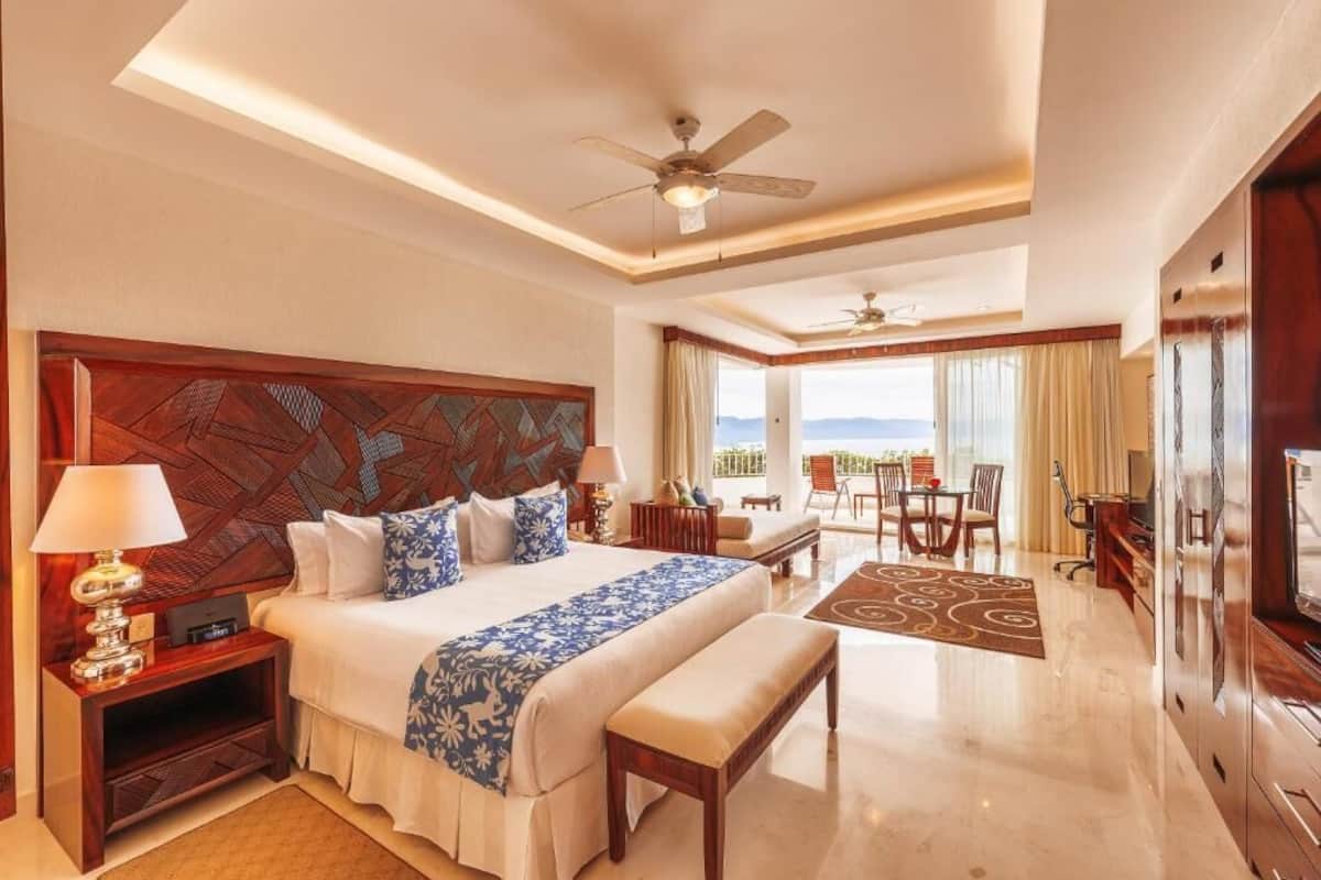 A hotel room with a bed, bedside tables, a desk, a chair, a TV, a ceiling fan, and a view of the outdoors.
