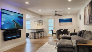 A modern living room with a flat-screen TV, a sofa, a dining area, and a kitchen. - Luxe Coastal Retreat • 12 Min to Beach • Private Yard w/Games (Clearwater)