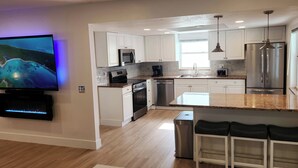 A modern kitchen with white cabinets, stainless steel appliances, a large island with bar stools, and a flat-screen TV mounted on the wall. - Luxe Coastal Retreat • 12 Min to Beach • Private Yard w/Games (Clearwater)