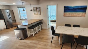 A modern kitchen with a stainless steel refrigerator, a granite countertop, and a dining area with a marble table and black chairs. - Luxe Coastal Retreat • 12 Min to Beach • Private Yard w/Games (Clearwater)