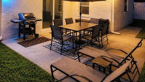 A patio with a grill, table, chairs, and an umbrella. - Luxe Coastal Retreat • 12 Min to Beach • Private Yard w/Games (Clearwater)