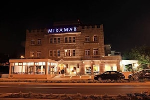 Um hotel chamado MIRAMAR com fachada de pedra, entrada de vidro moderna e área de estacionamento com carros.