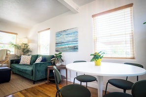 A living room with a green sofa, a round white table, and a painting of a beach scene. - Charming 1BR bungalow in sunny Ocean Beach with yard (San Diego)
