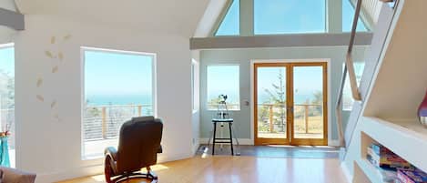Une pièce spacieuse avec un plafond haut, un fauteuil à bascule, un télescope et un balcon offrant une vue sur l’océan.