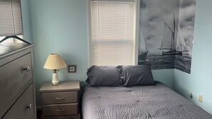 A bedroom with a bed, a nightstand, a lamp, a dresser, a window with blinds, and a wall with a seascape painting.