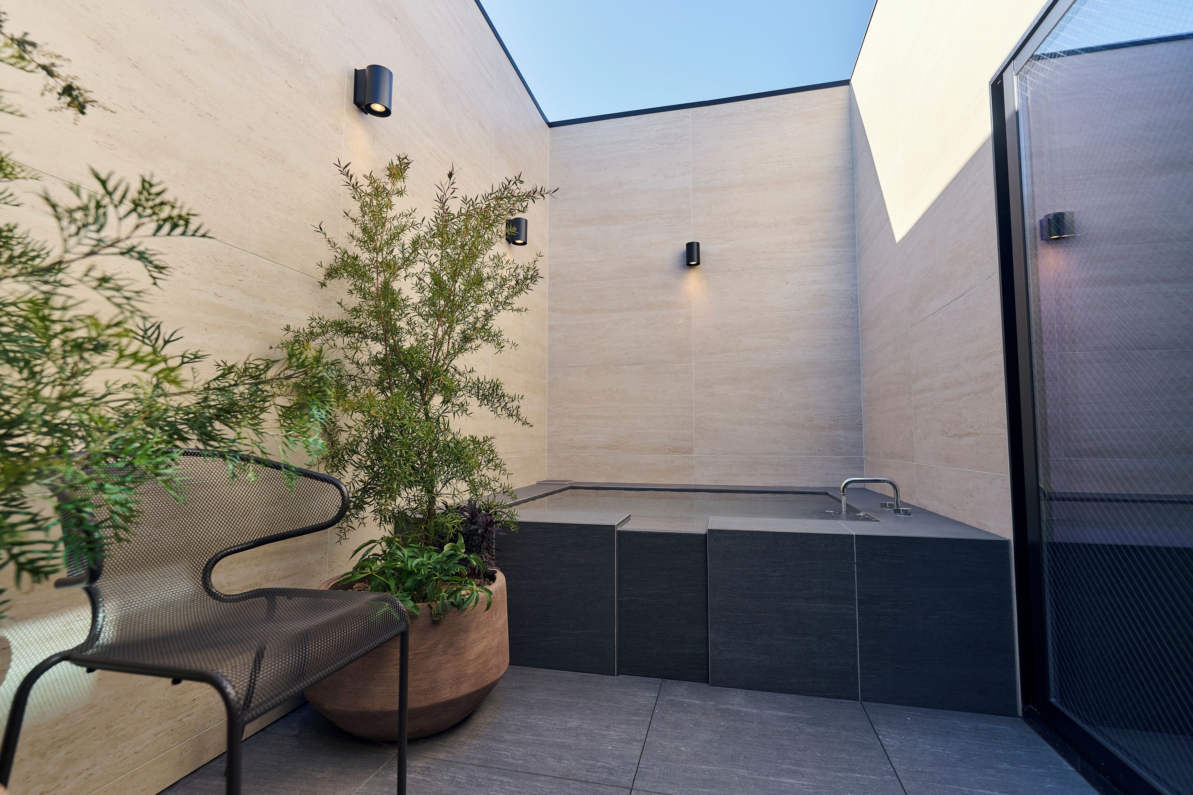 Une salle de bain extérieure moderne dotée d’une baignoire intégrée, d’un espace détente avec un banc et des plantes en pot, ainsi que d’un luminaire fixé au mur.
