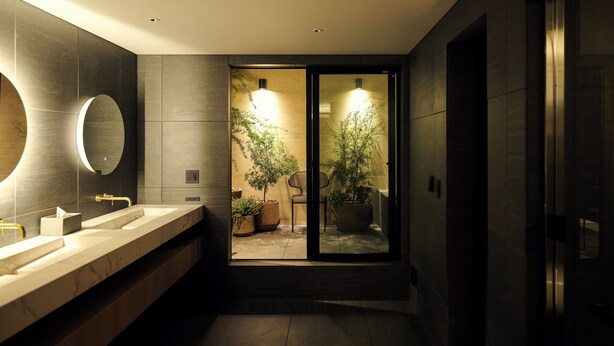 A modern bathroom with a large mirror, two sinks, and a view into a room with plants and a chair.
