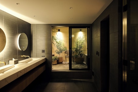 A modern bathroom with a large mirror, two sinks, and a view into a room with plants and a chair.