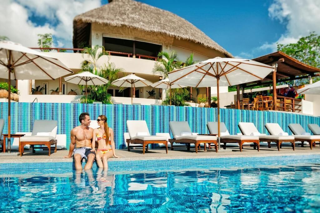 Una pareja junto a una piscina con sillas de descanso y sombrillas, un edificio con techo de paja y un cielo azul despejado.
