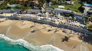 Vista aérea de un complejo turístico en la playa, con una extensa playa de arena, una zona de piscina con sombrillas y algunas palmeras.