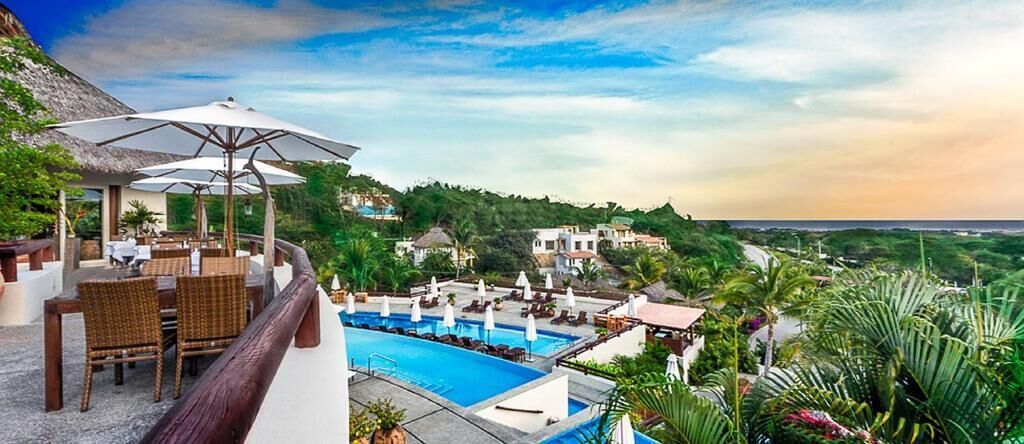 Ein Poolbereich mit Liegestühlen und Sonnenschirmen, mit freiem Blick auf den Pool, und eine Hügelkuppe mit Gebäuden im Hintergrund.