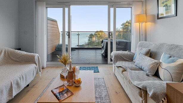 A living room with a sofa, a coffee table with a book and a bottle, and a view of the sea through a glass door. - Avocet 2 is a stylish coastal apartment offering uninterrupted sea views (Brixham)