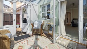 Un espace de vie lumineux et décloisonné, avec une porte-fenêtre coulissante donnant sur une chambre, des meubles en osier et une petite table avec une lampe.